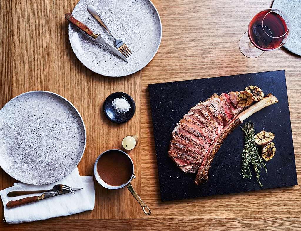 Sofitel Sydney Darling Harbour Hotel Exterior photo The photo features a beautifully arranged dining setup. At the center, there is a large, cooked rib steak presented on a black serving board, garnished with herbs and roasted garlic. Surrounding the steak are round, textured plates, each paired with 
