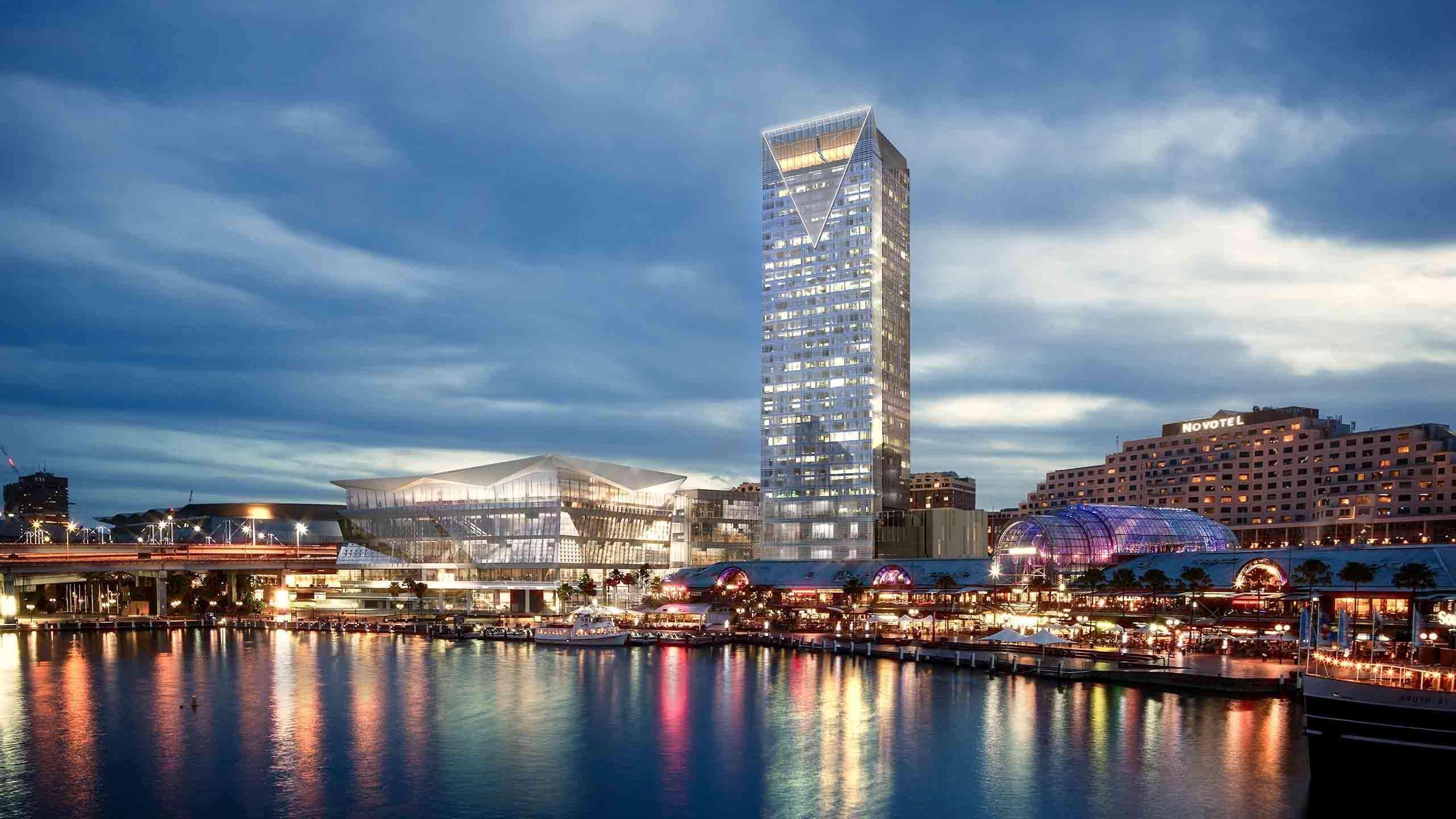 Sofitel Sydney Darling Harbour Hotel Exterior photo The photo features a modern, illuminated skyscraper next to a body of water during twilight. The building has a sleek design with a pointed top and large glass windows that reflect the surrounding light. In the foreground, there are colorful lights f
