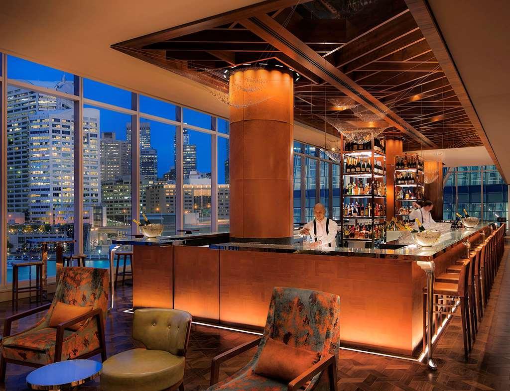 Sofitel Sydney Darling Harbour Hotel Exterior photo The photo depicts an upscale bar located in a modern setting. It features a sleek design with large windows that offer a view of a city skyline, illuminated by twilight. The bar itself is well-lit and showcases a variety of bottles on shelves behind 