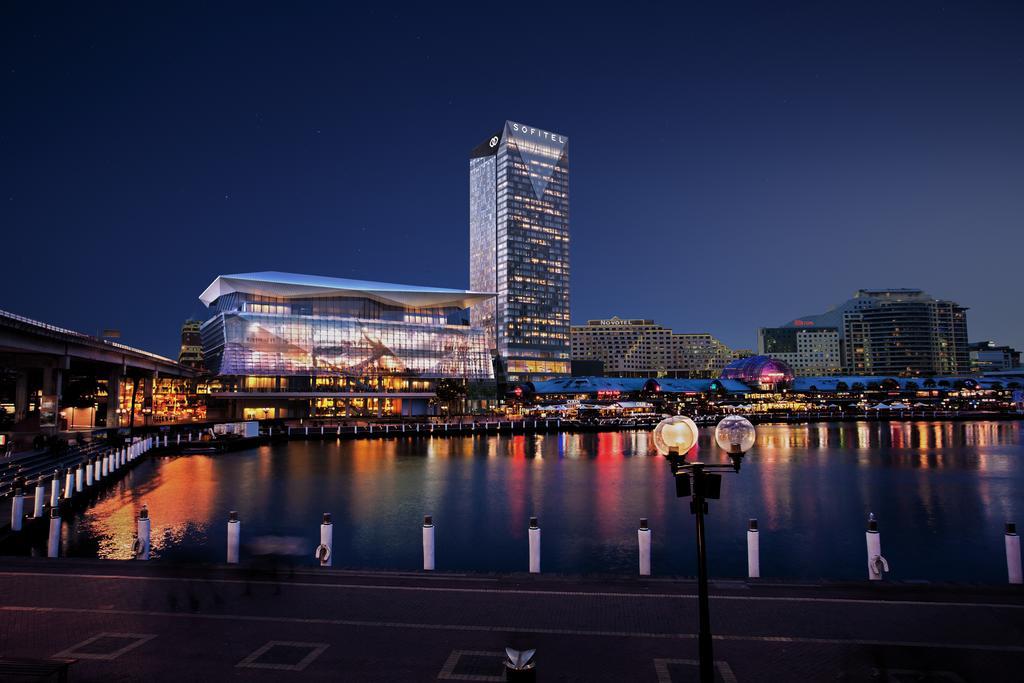 Sofitel Sydney Darling Harbour Hotel Exterior photo The photo depicts a nighttime scene featuring a modern waterfront area. In the foreground, there is a body of water reflecting the lights from surrounding buildings. Prominent in the background is a tall, sleek skyscraper, which is illuminated and st