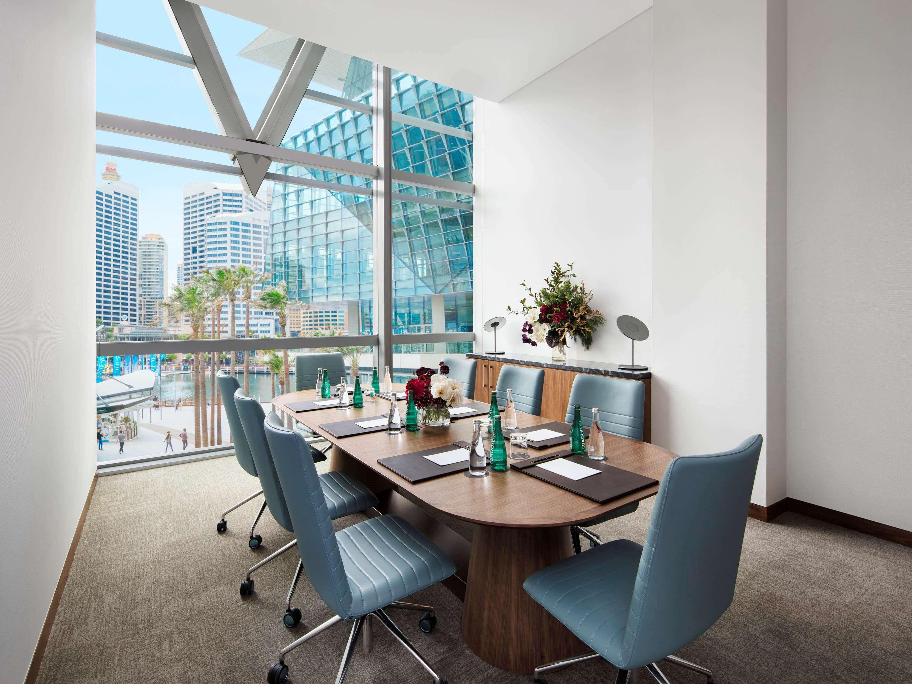 Sofitel Sydney Darling Harbour Hotel Exterior photo The photo shows a modern conference room featuring a large wooden table surrounded by several blue chairs. Large windows provide a view of a cityscape with tall buildings and greenery outside. The room is well-lit with natural light, and there are de