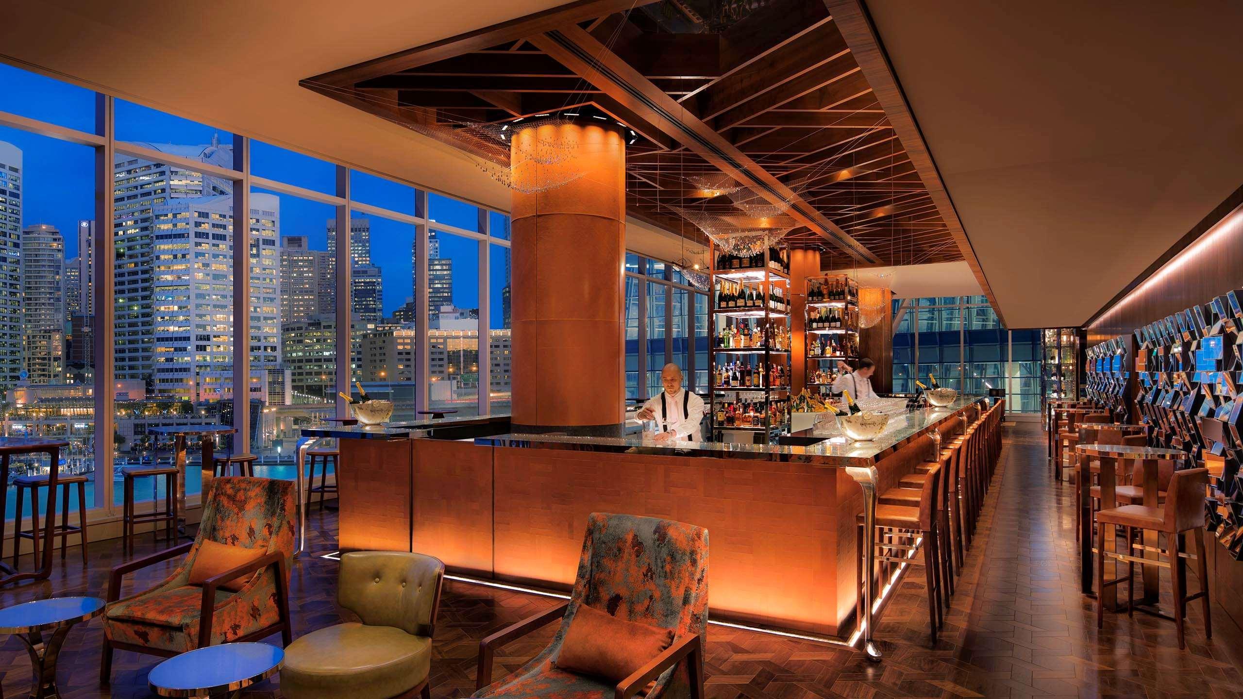 Sofitel Sydney Darling Harbour Hotel Exterior photo The photo shows a stylish bar interior, likely located in a high-rise building given the large windows that reveal a city skyline at dusk. The bar features a modern design with warm lighting, wooden accents, and a polished countertop. Behind the bar,