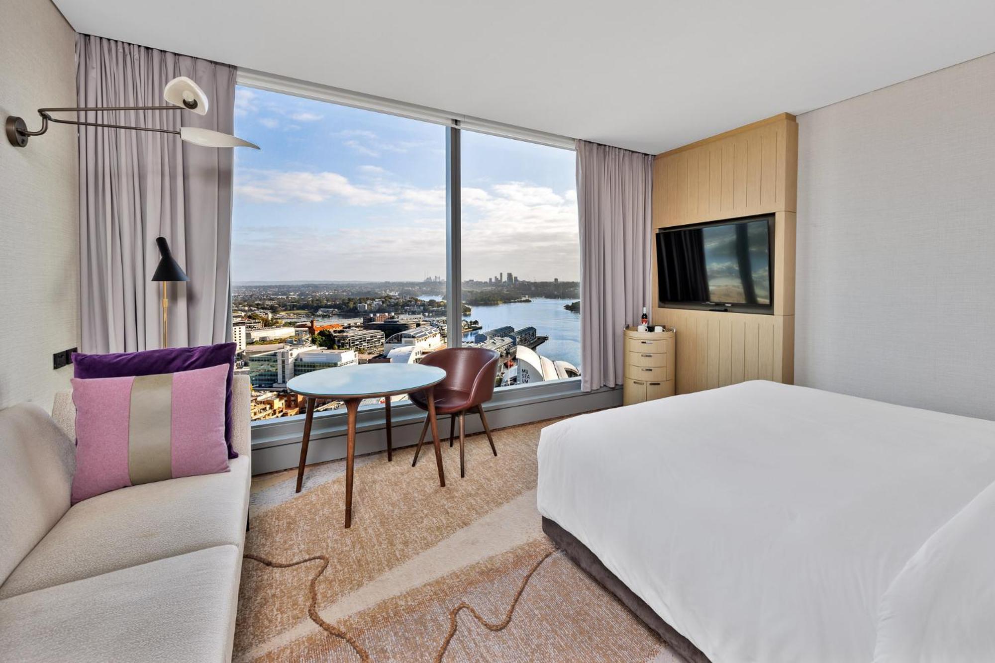 Sofitel Sydney Darling Harbour Hotel Exterior photo The image shows a modern hotel room featuring a large window with a scenic view. Inside the room, there is a bed with white linens, a cozy sofa with colorful cushions, and a small round table with two chairs. A flat-screen TV is mounted on the wall, 