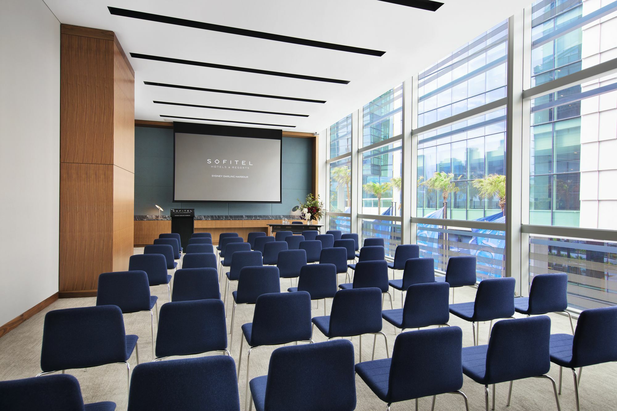 Sofitel Sydney Darling Harbour Hotel Exterior photo The photo shows a modern conference room or meeting space. It features rows of blue chairs arranged in front of a presentation screen mounted on a wall. The room has large windows, allowing natural light to enter, and offers a view of an outdoor area