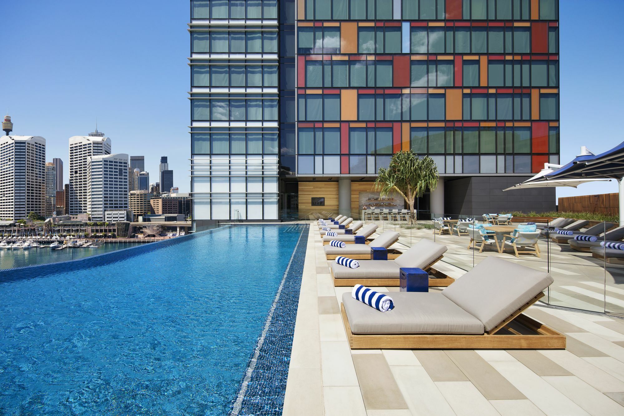 Sofitel Sydney Darling Harbour Hotel Exterior photo The photo shows a modern, upscale pool area on the rooftop of a building. There is a large infinity pool with clear blue water extending towards the edge of the image, creating a seamless view with the skyline in the background. Alongside the pool, t