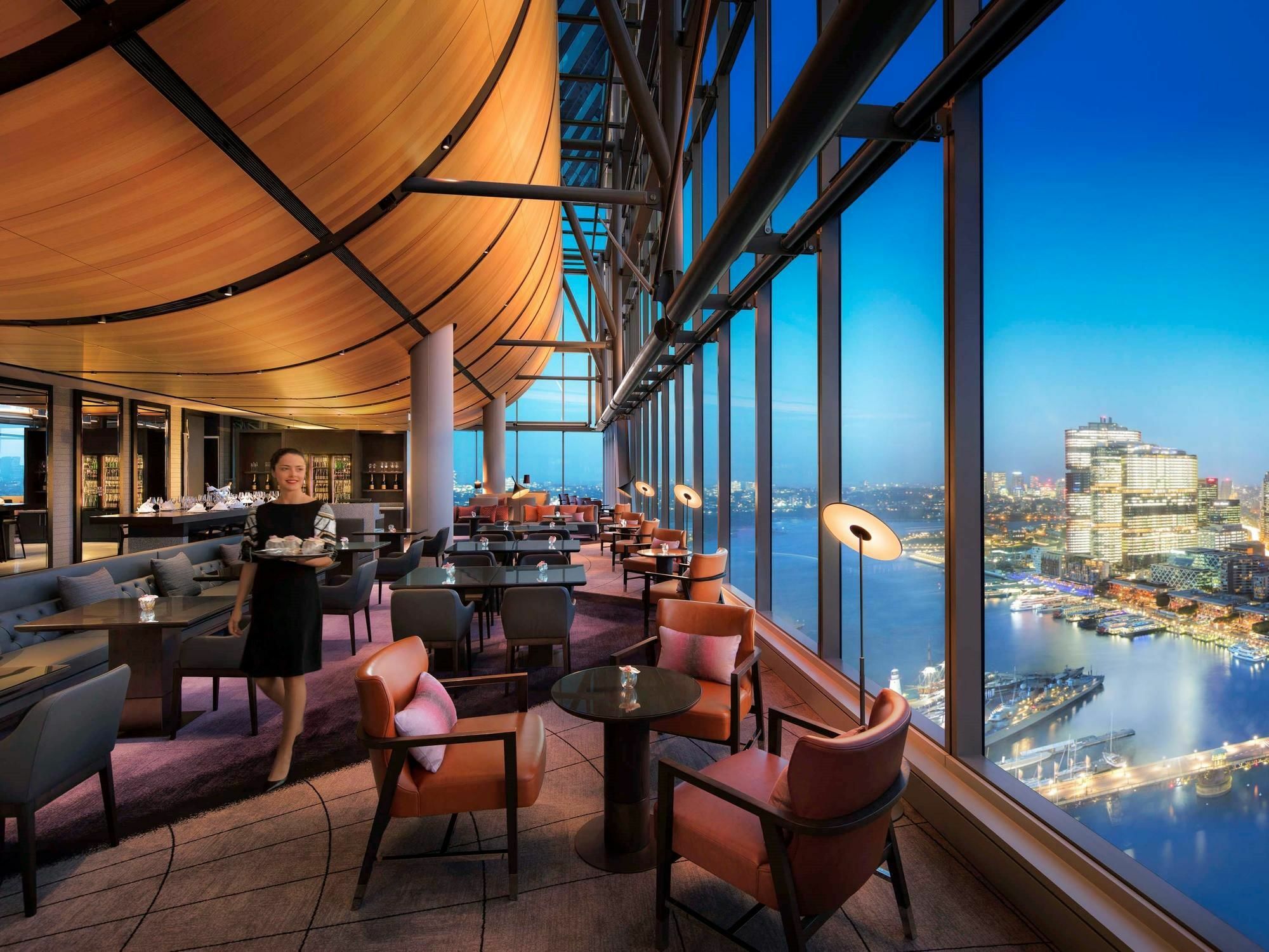 Sofitel Sydney Darling Harbour Hotel Exterior photo The photo depicts an elegant dining area located in a high-rise building, featuring a panoramic view of a city skyline during twilight. The interior design includes modern furnishings with comfortable chairs, polished tables, and large windows that a